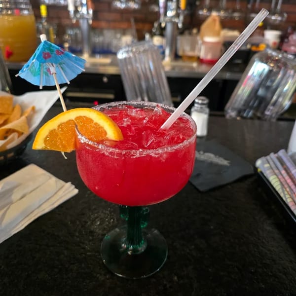 A vibrant red margarita with an orange slice and blue umbrella sits on a bar.