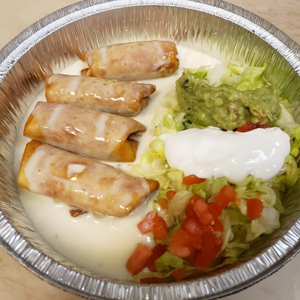 Four taquitos covered in white sauce, with guacamole, sour cream, lettuce, and diced tomatoes in a foil container.