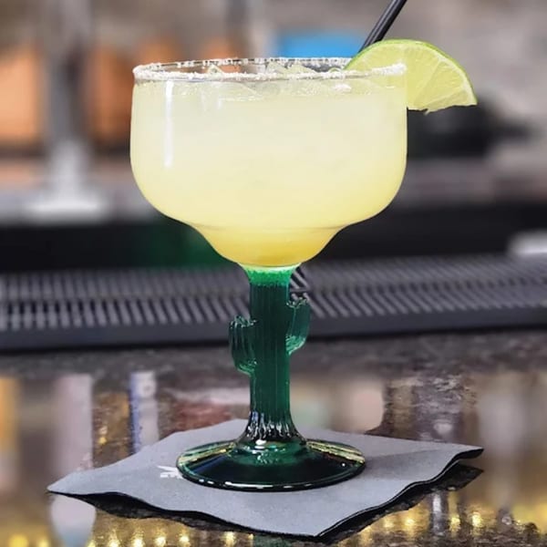 Margarita in a cactus-stemmed glass with a salted rim and lime wedge on a dark napkin.