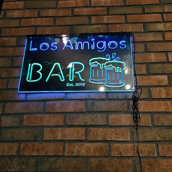 Neon sign for Los Amigos Bar, established 2013, with two beer mugs, on a brick wall.