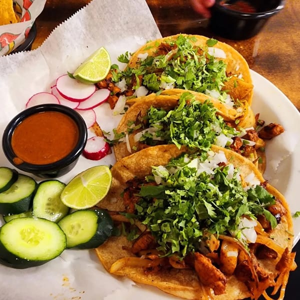Three Los Amigos tacos with cilantro, onions, lime, radishes, cucumbers, and a side of salsa.